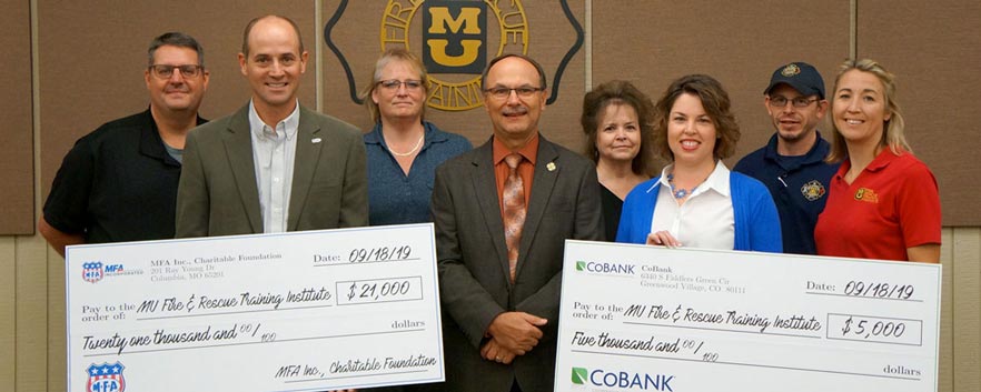 Eight people standing in front of a wall with the MU Fire and Rescue Training Institute's emblem while oversized checks for $21,000 from MFA and $5,000 from CoBank are being presented to the organization.