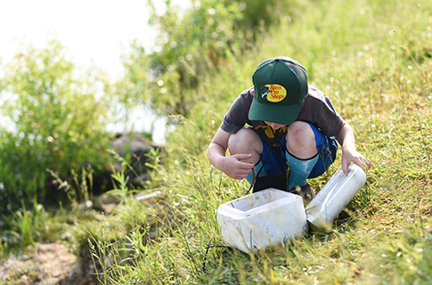 A boy looking into a foam cooler near the water of a lake.