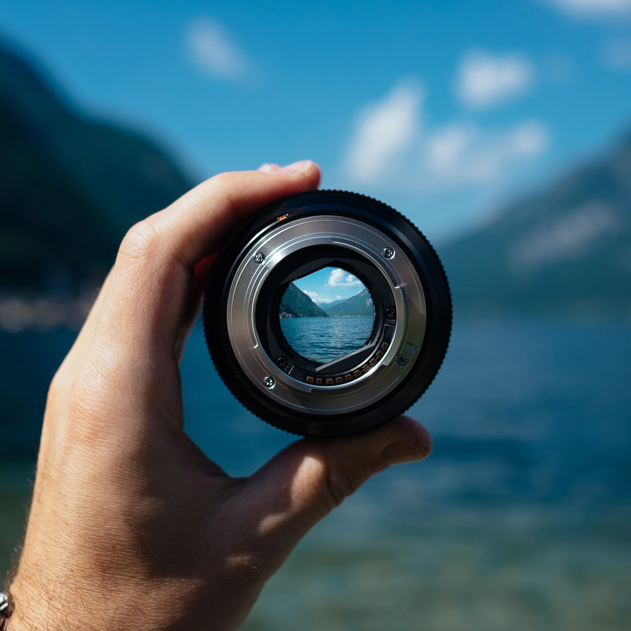 A hand holds a camera lens up in front of a moutain and lake view.
