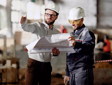 Two men wearing safety hats discussing blue prints plans.