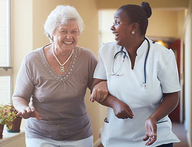 An elderly woman and a female nurse laugh as they walk arm and arm down a hallway.