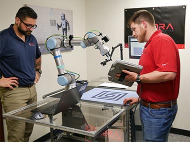 Adam Haney and Colby Thrift workking at a clear table with a laptop and a robotic arm on it. Photo property of Mora Technologies. Used with permission.