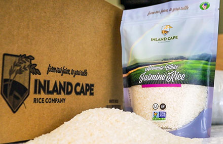 A pile of rice in front of a bag of Inland Cape Rice beside a branded box it is shipped in.