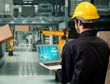 Supply chain manager using laptop in warehouse where robotic arms are moving large shipping boxes