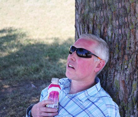 Man laying against tree with sun burnt face, sunglasses and plastic bottle in hand.