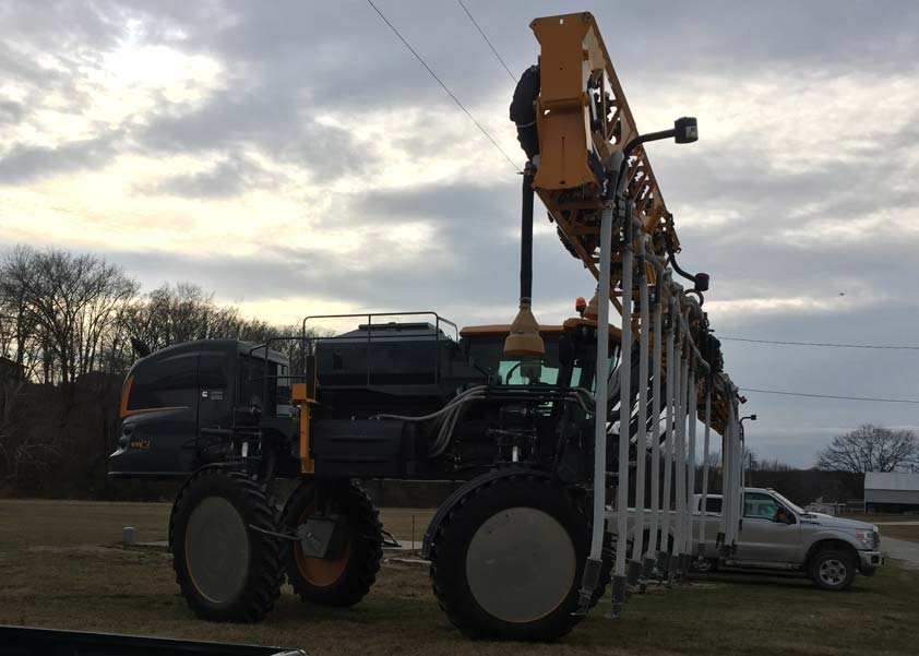 A high-clearance sprayer equipped with seeding equipment in a growing corn field.