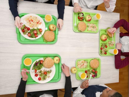 Aerial view of a five people sitting at a cafeteria table eating a healthy lunch from green trays.