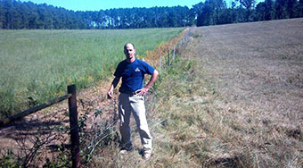 Man standing next to a barbed wire fence with a green field to the left of the fence, and a brown field to the right of the fence.