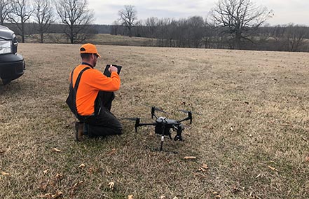 A technician reviews footage from a drone and prepares for surveillance assistance using the drone next to him for aerial operations for feral hog removal.