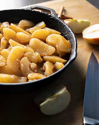 An iron pot of fried apples on a cutting board beside a knife and some sliced applies.