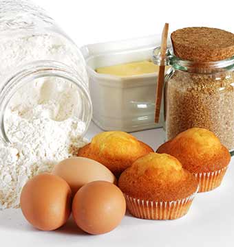Homemade muffins beside some eggs, a dish of butter and a jar of flour.