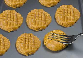 A pan of peanut butter cookies with crisscrossed fork pattern; one is being pressed by a fork.