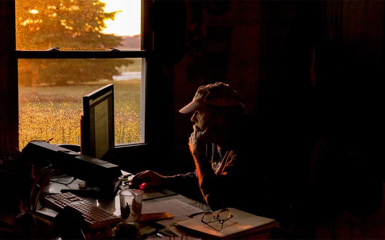 Man in a ball cap sits at a computer as dim orange light enters the room through a nearby window.