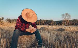 Man in field sitting on hay bale with his head hidden under a hat.