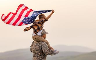 Soldier carrying a boy on his shoulders who is gripping an American flag flapping in the breeze.