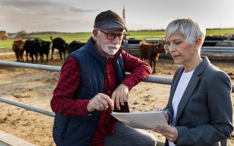 Man leans on the fence to a corral and looks at papers held by a woman.