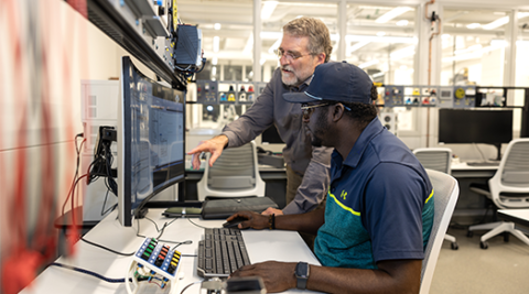 individual seated at computer. Another individual standing, leaning in toward seated person and pointing to screen