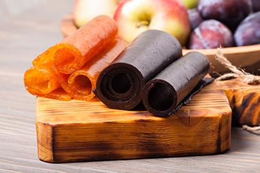 Dehydrated fruit leathers on a cutting board with a bowl of fruit behind them