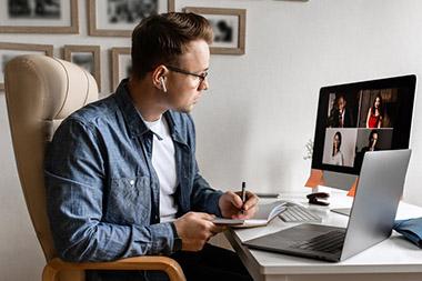 Man sitting at desk in home office on virtual meeting with participants pictured on his monitor.