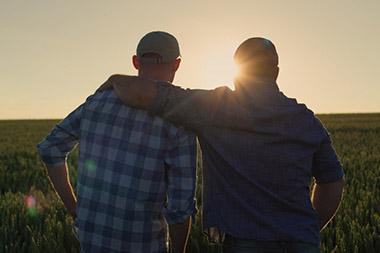 Father with arm around adult son as they stand in a field watching the sunset.