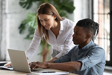 Woman standing over man's shoulder as they look at his laptop screen.