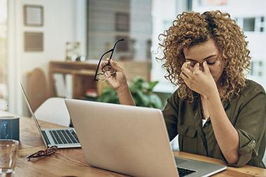Woman sitting at table with 2 open laptops looking exasperated as she has removed her eyeglasses and is rubbing her eyes.
