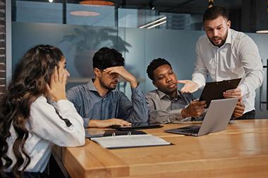 Man showing clipboard to three people sitting at table in conference room and everyone looks tense.