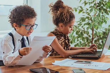 Young children in workplace attire in an office setting with a boy reading a report and a girl typing on a laptop.