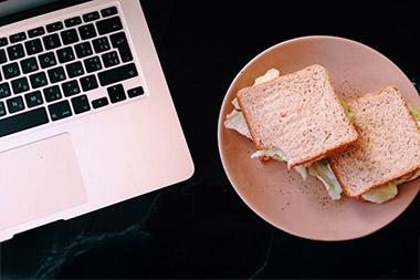A tabletop with an open laptop next to a sandwich on a plate