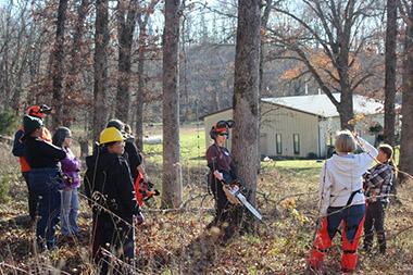 Participants in woods watching a woman teach chainsaw safety