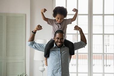 Young boy sitting on dad's shoulders as they both flex their arm muscles.