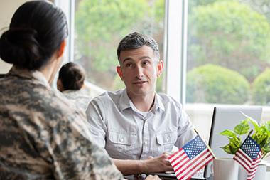 A man and woman speaking at a table with American flags on it.
