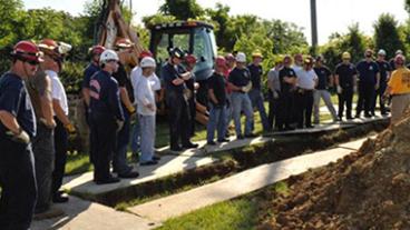 A group wearing hard hats standing over a trench.