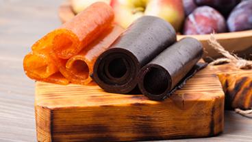Dehydrated fruit leathers on a cutting board with a bowl of fruit behind them