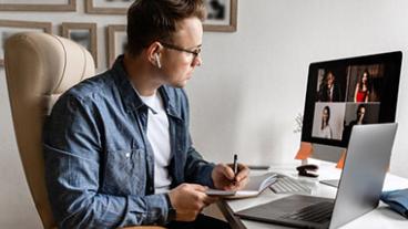 Man sitting at desk in home office on virtual meeting with participants pictured on his monitor.