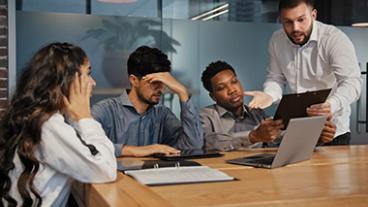 Man showing clipboard to three people sitting at table in conference room and everyone looks tense.