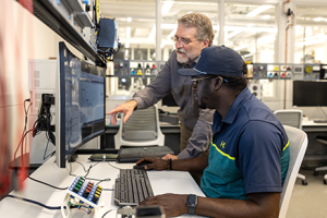 individual seated at computer. Another individual standing, leaning in toward seated person and pointing to screen