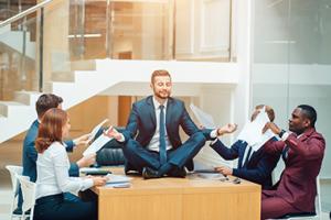 Man sitting on desk meditating while co-workers exchange ideas