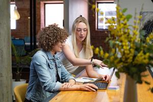 Two women sitting at a table talking while typing something on a laptop.
