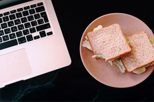 A tabletop with an open laptop next to a sandwich on a plate