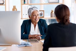 Woman in leadership role smiling and talking to another woman sitting on the other side of her desk. 