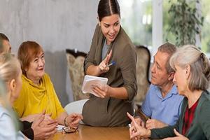 A group of seniors at a table talking with a woman holding a notebook.