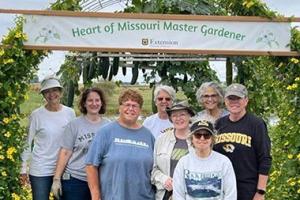 Heart of Missouri Master Gardeners standing under a flower trellis and the HMMG banner.